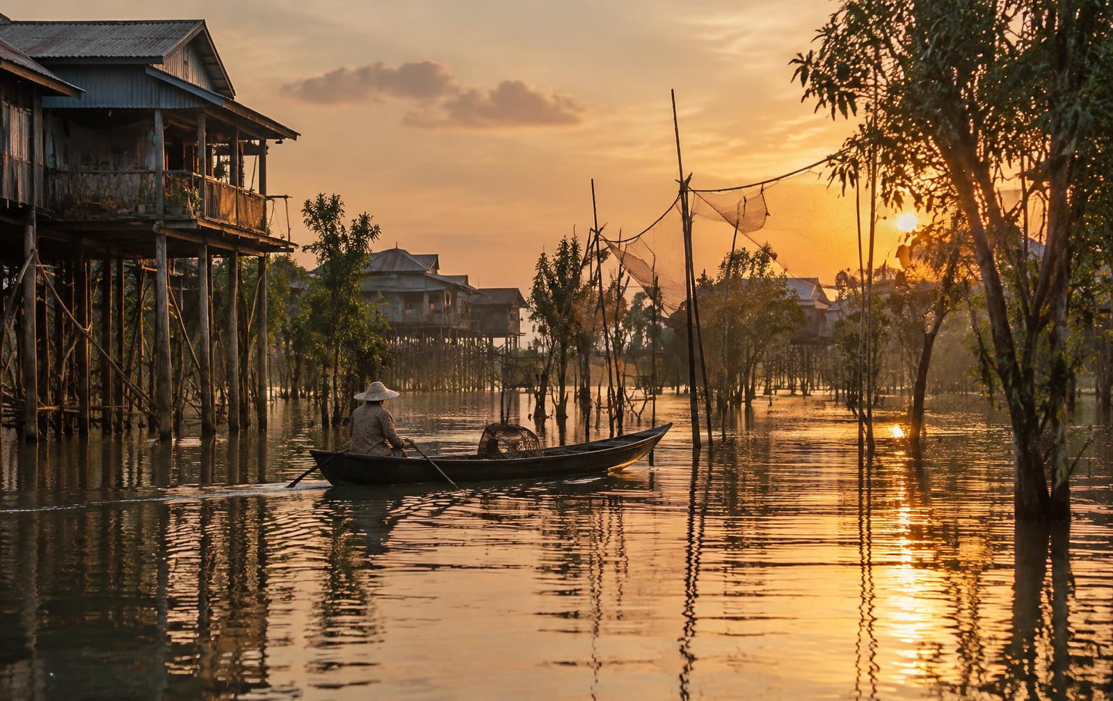 Stilted wooden houses and a narrow sampan boat gliding through the calm golden waterways of Kampong Khleang on Tonlé Sap Lake near Siem Reap